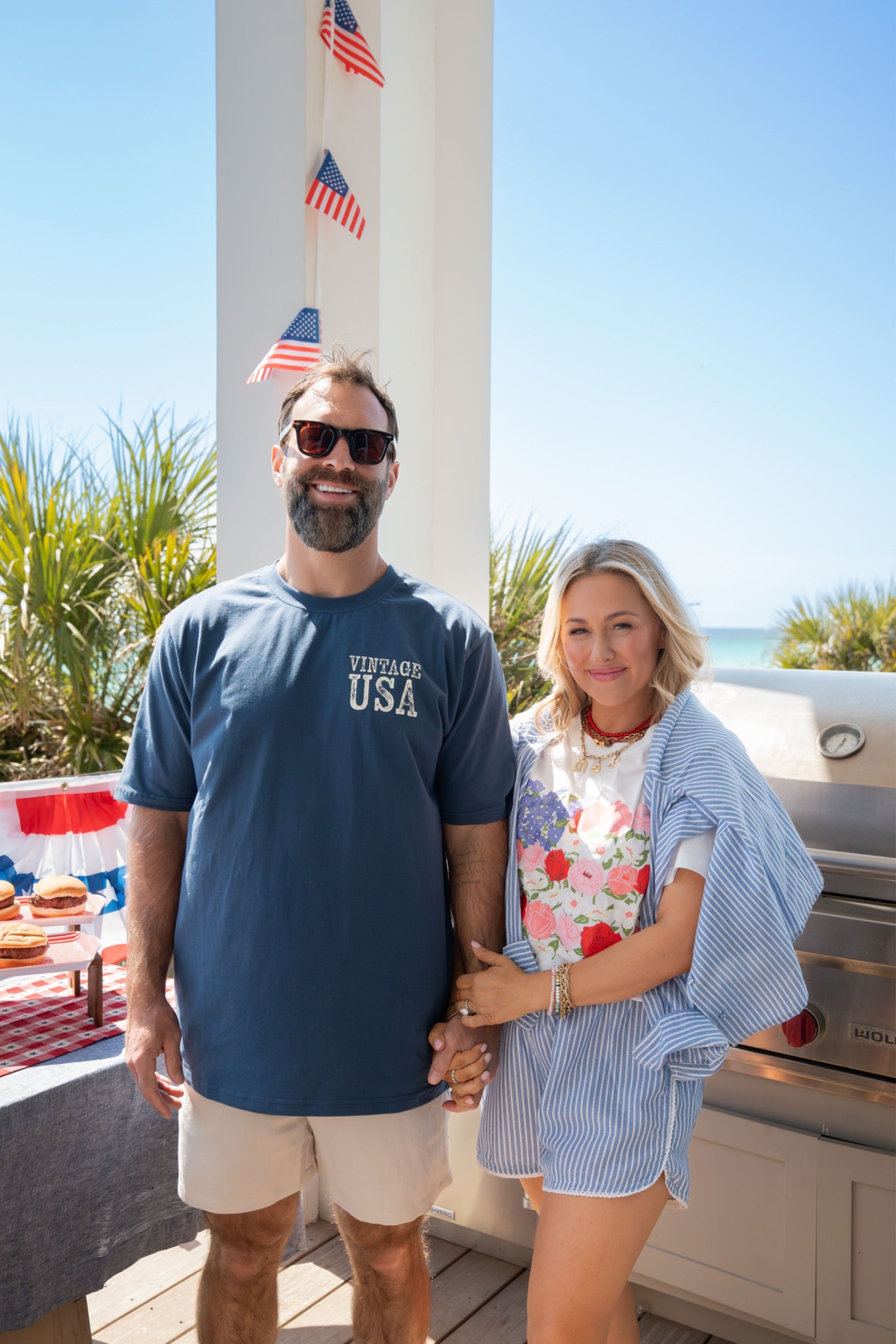 Man and woman standing outdoors with American flags and a barbecue in the background.
