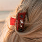 Close-up of a red hair clip with decorative elements in blonde hair against a blurred natural background.