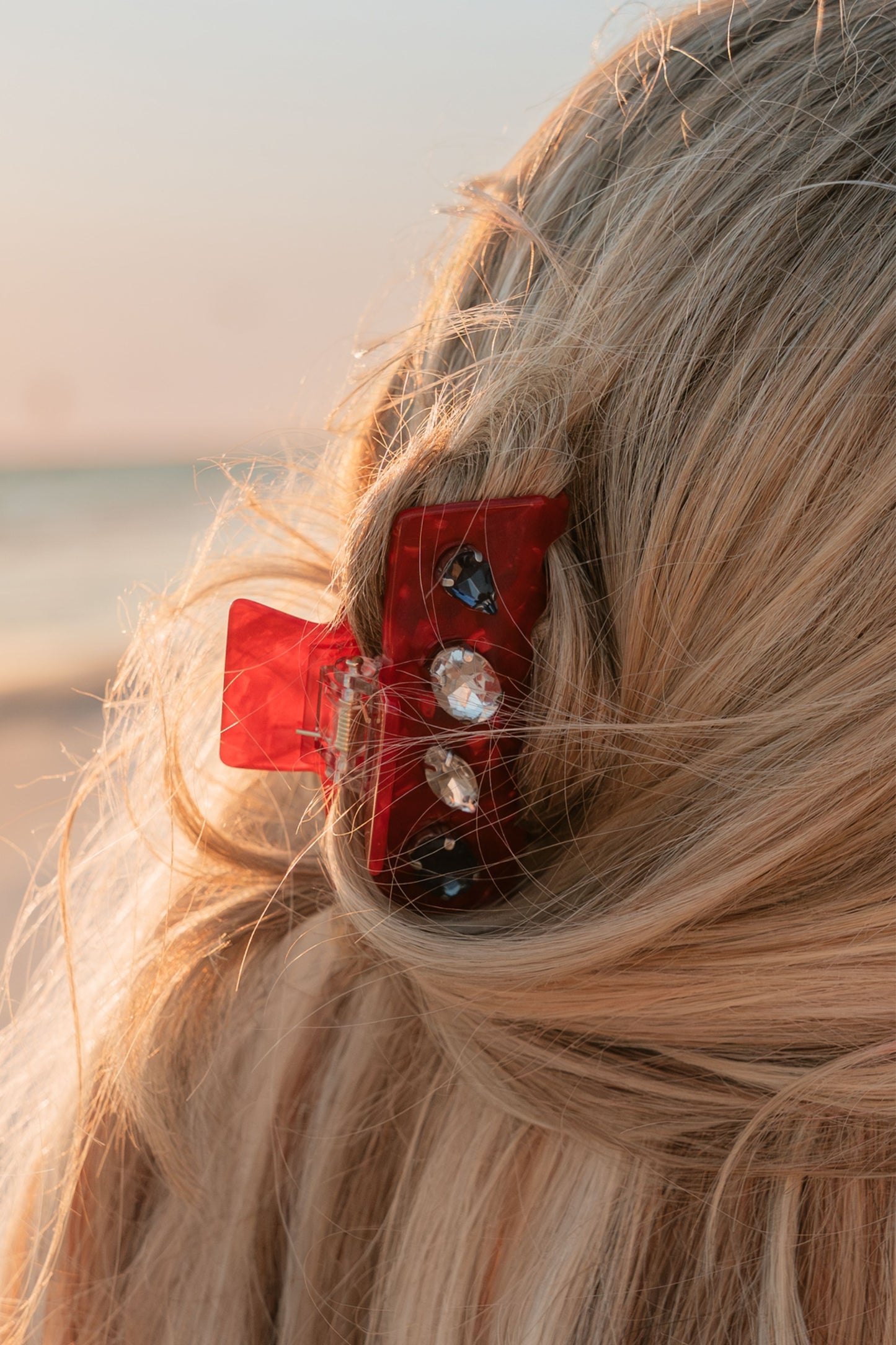 Close-up of a red hair clip with decorative elements in blonde hair against a blurred natural background.