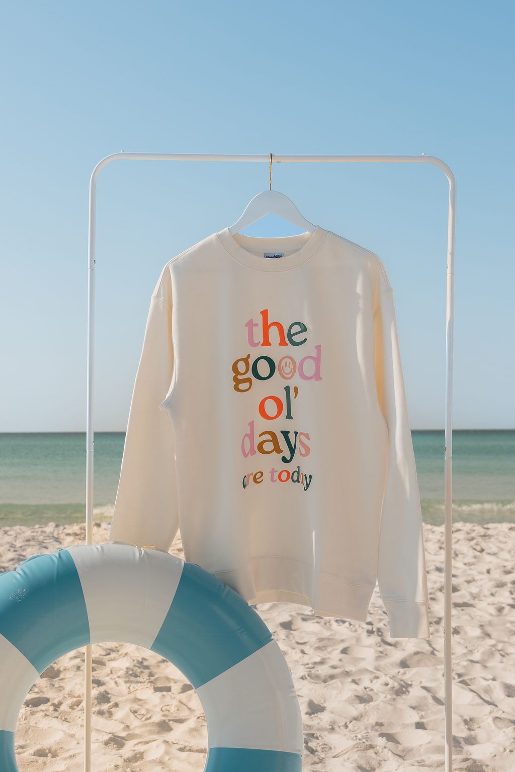 Beige sweatshirt with colorful text on a beach with a blue sky and ocean.