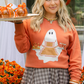 Woman in an orange sweater with ghost and pumpkin design, holding a tray of candy apples.