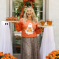 Woman in an orange sweatshirt with ghost design holding drinks, standing in front of a decorated door with pumpkins and flowers.