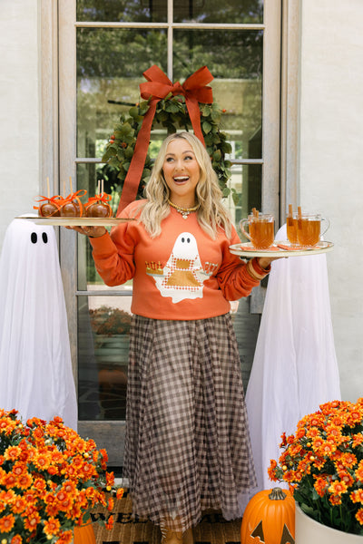 Woman in an orange sweatshirt with ghost design holding drinks, standing in front of a decorated door with pumpkins and flowers.