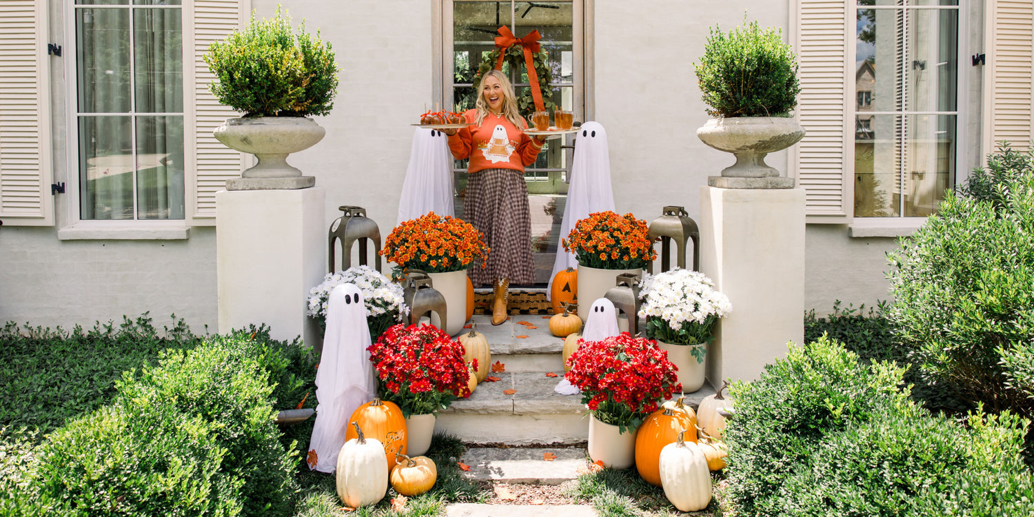 Woman standing on a decorated front porch with Halloween-themed decorations including pumpkins, ghosts, and flowers. She is wearing a ghost sweatshirt.