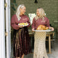 Two women in matching sweatshirts holding platters of food in a home setting.
