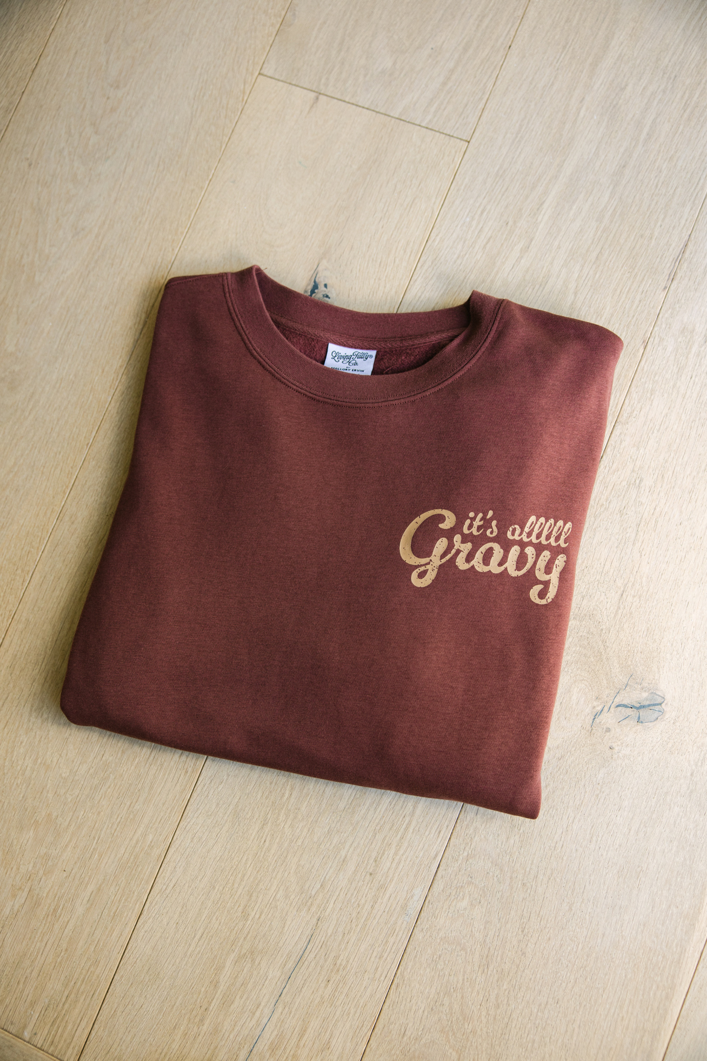 Folded brown sweatshirt with 'it's alllll Gravy' text on a wooden floor.