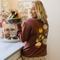 Woman in a kitchen holding a bowl, wearing a brown sweater with food-themed designs.