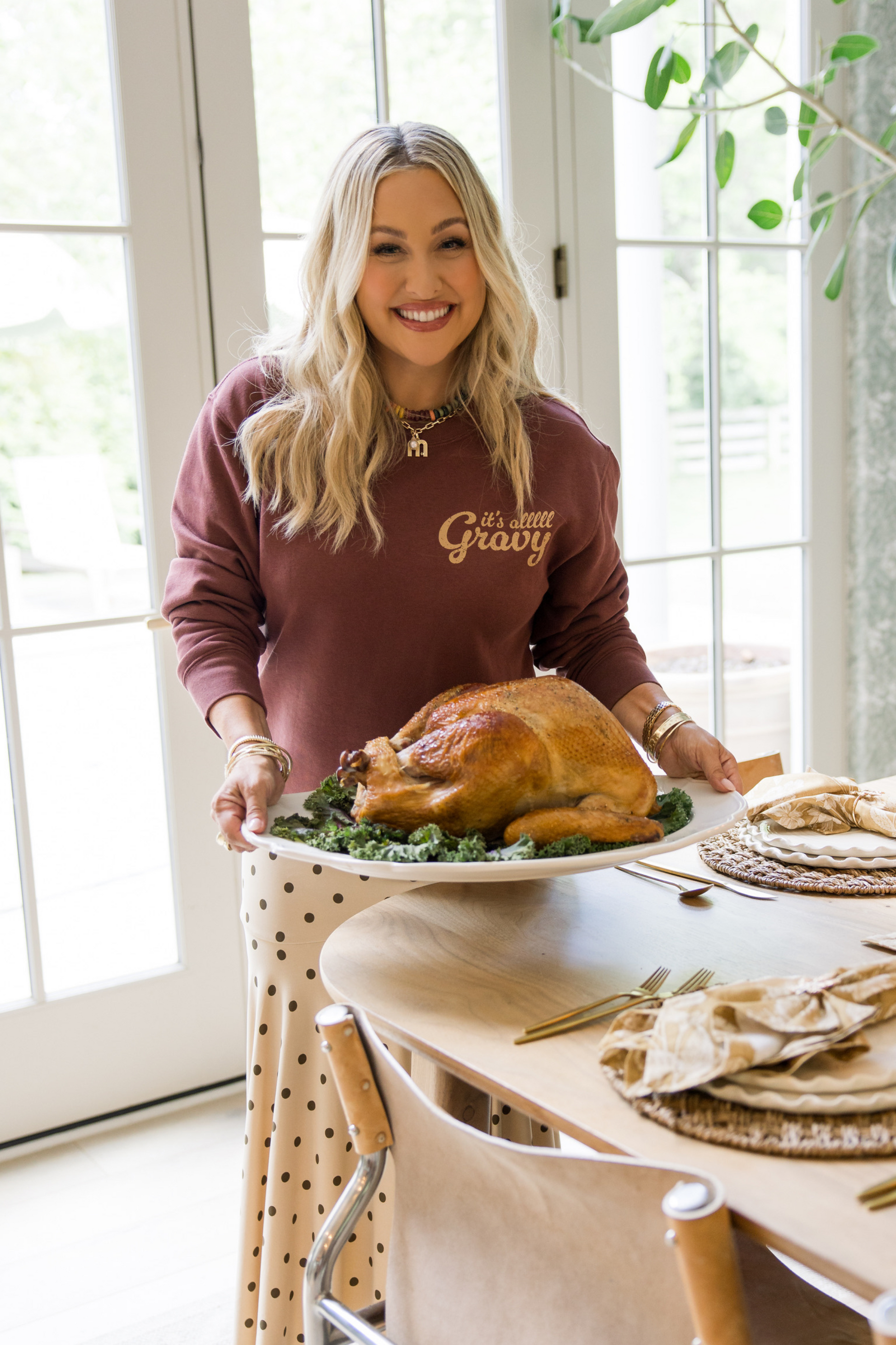 Woman in a maroon sweatshirt with 'it's just gravy' text, holding a roasted turkey on a dining table.