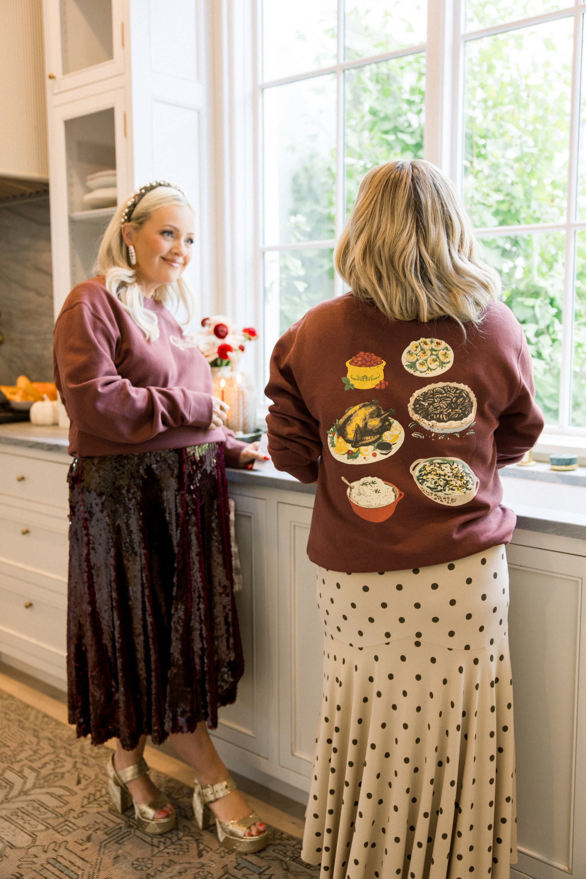 Two women in a kitchen, one wearing a sweater with food graphics.
