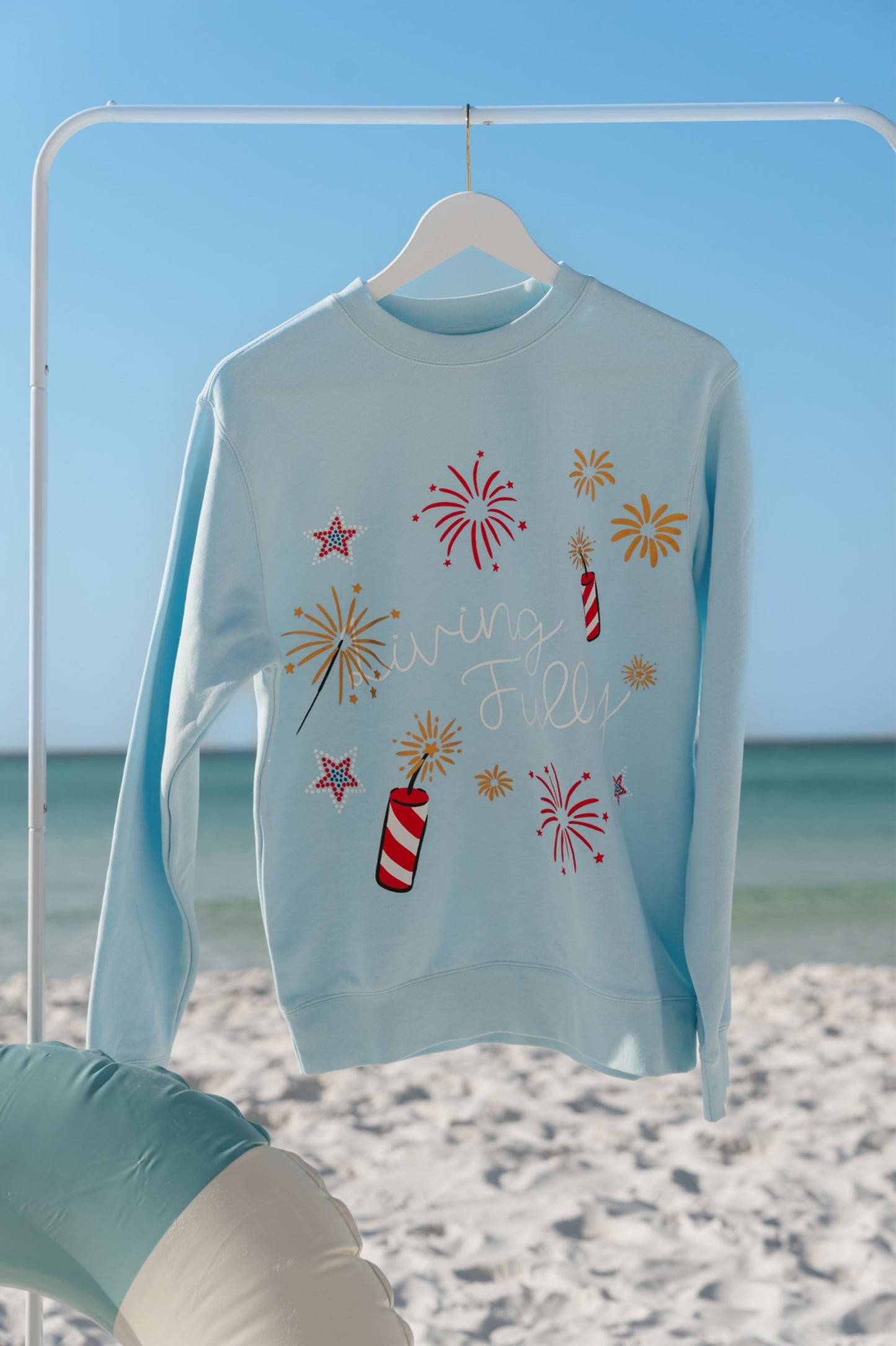 Light blue sweatshirt with festive designs on a hanger against a beach backdrop with a firework design and "Living Fully" text.