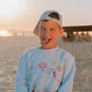 Child wearing a light blue LFC kids sweatshirt with colorful graphics and text, sitting on a beach.