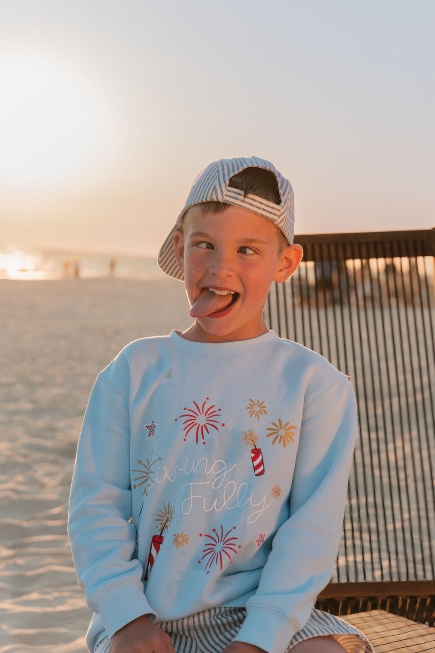 Child wearing a light blue LFC kids sweatshirt with colorful graphics and text, sitting on a beach.