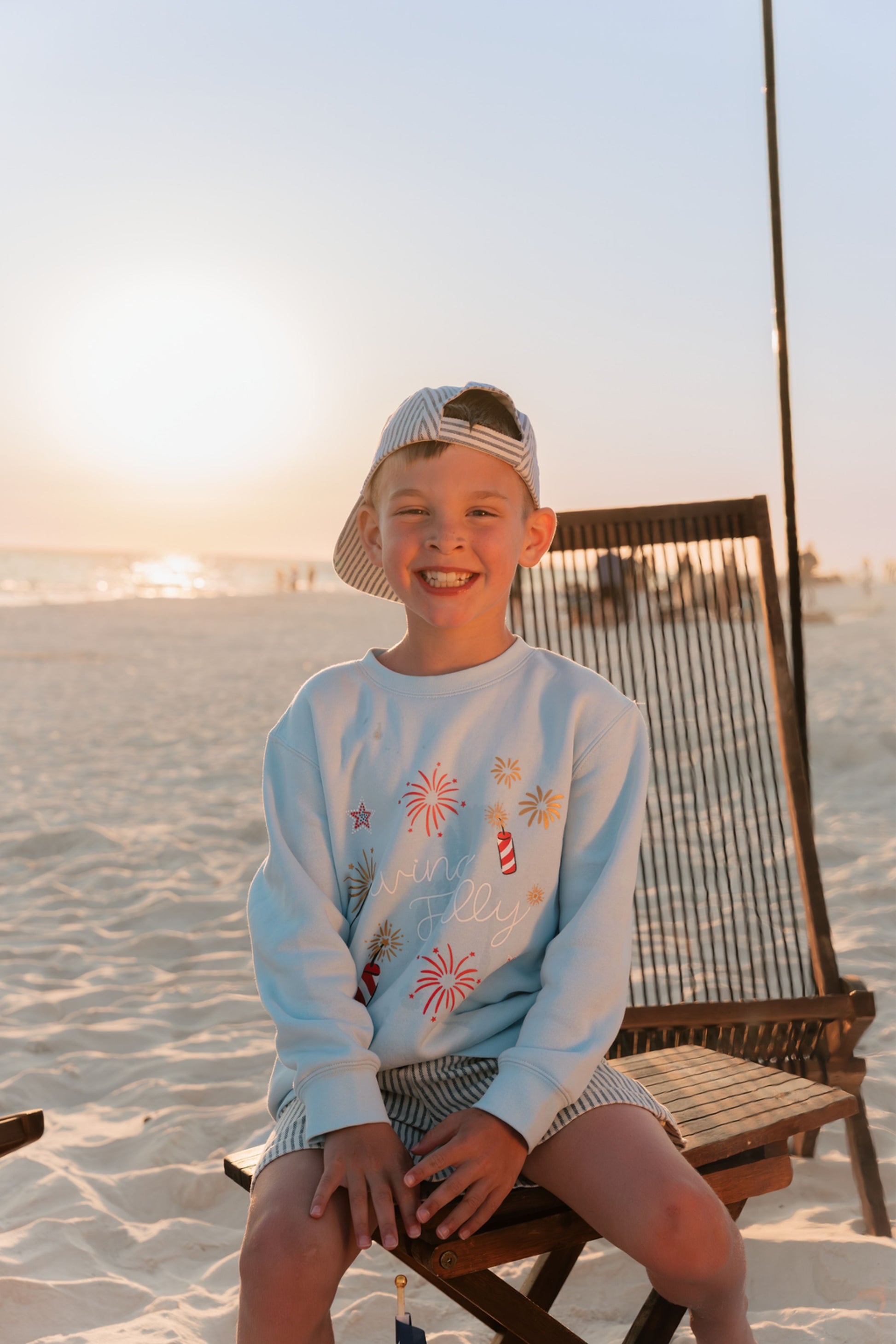 Child sitting on a beach chair at sunset with a light blue LFC kids sweatshirt and cap.