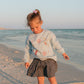 Young girl standing on a beach at sunset wearing a light blue LFC kids sweatshirt with firework designs and a patterned skirt.