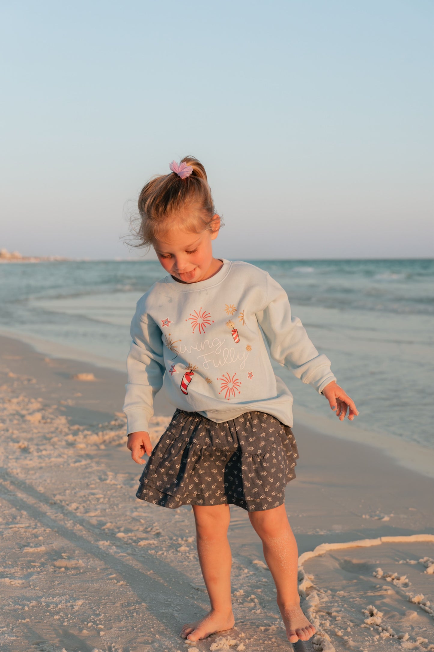 Young girl standing on a beach at sunset wearing a light blue LFC kids sweatshirt with firework designs and a patterned skirt.