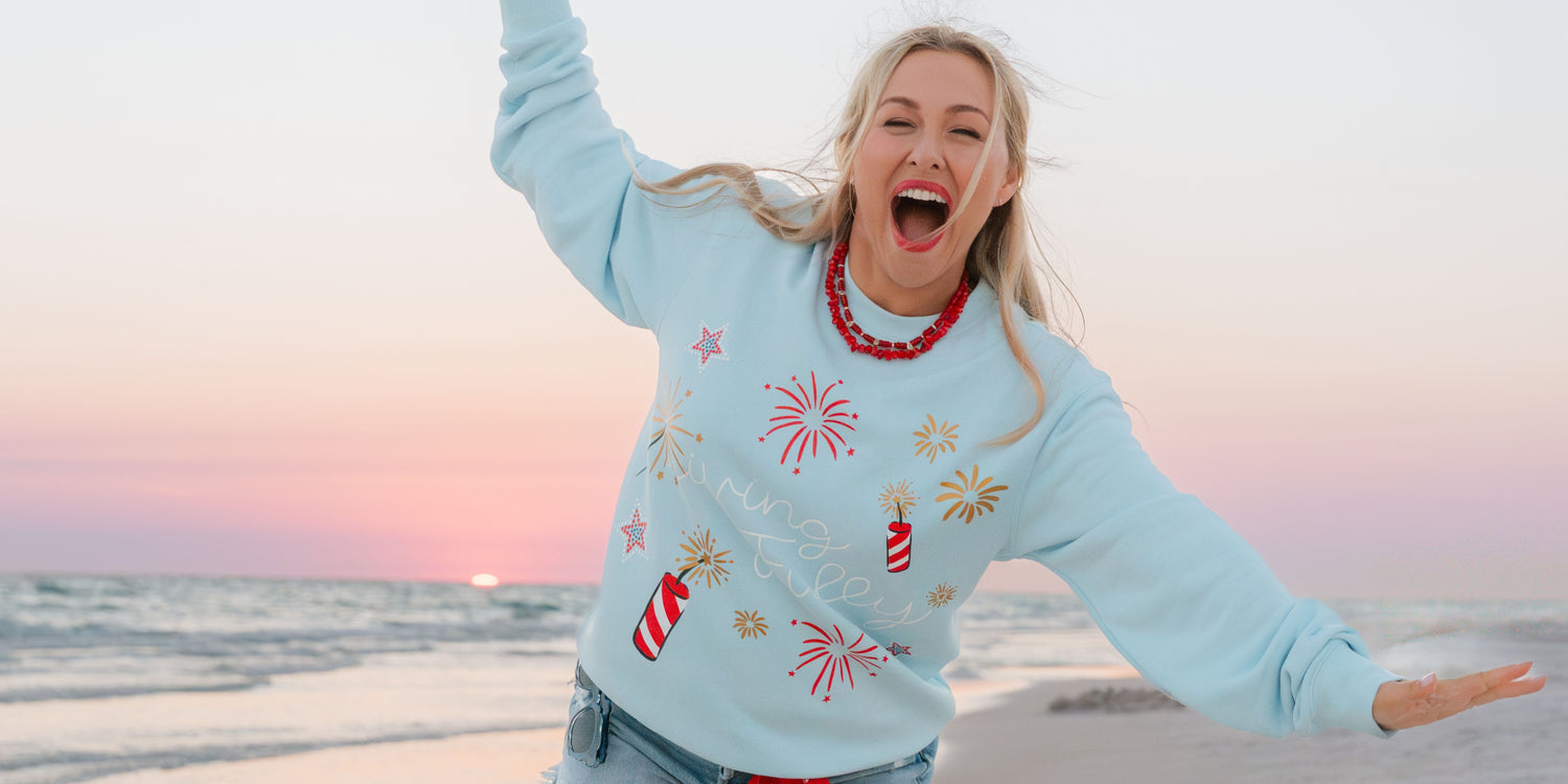 Woman wearing a festive Independence Say sweatshirt on a beach at sunset.