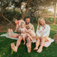 Family of five sitting on a blanket in a park with trees and grass.
