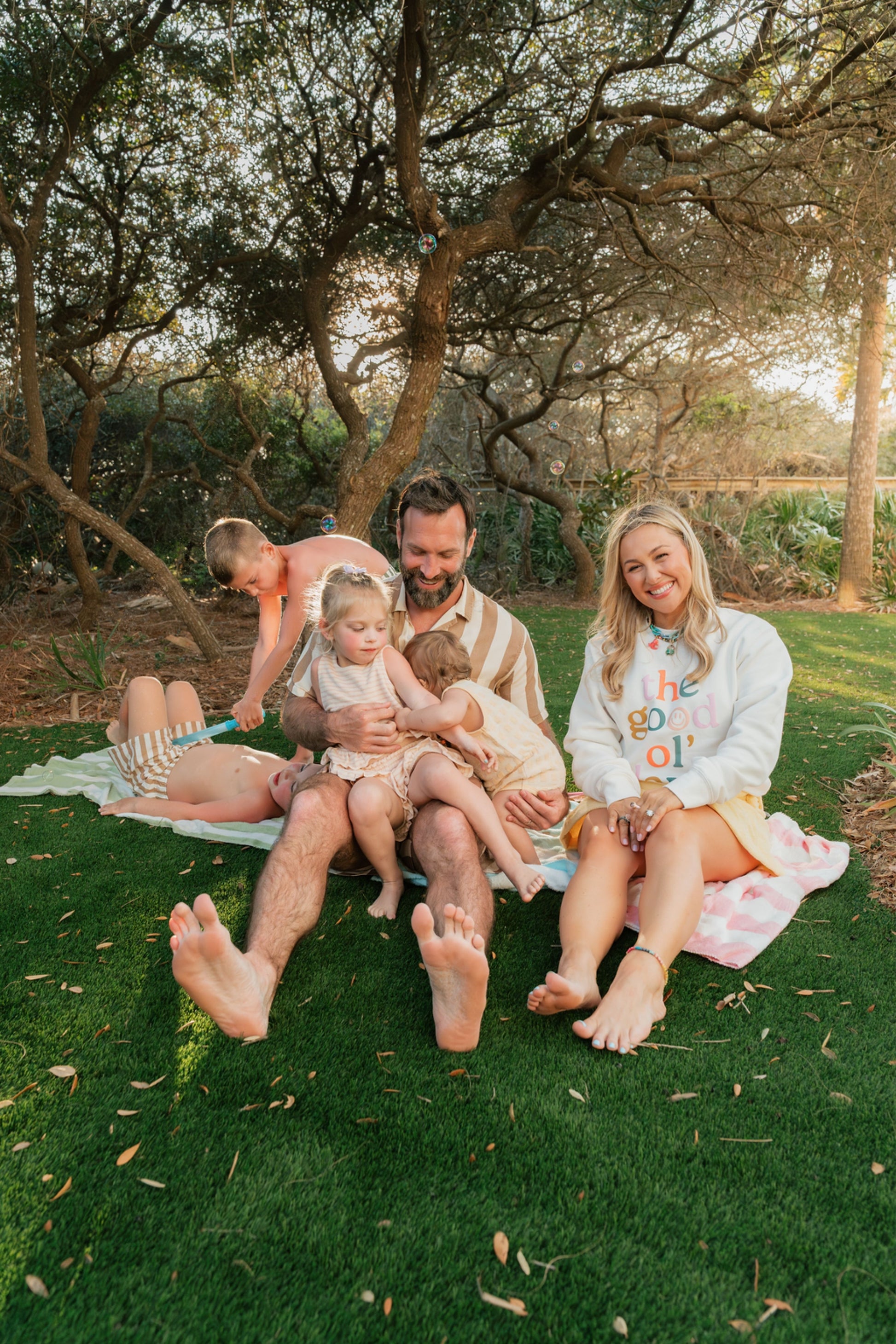 Family of five sitting on a blanket in a park with trees and grass.