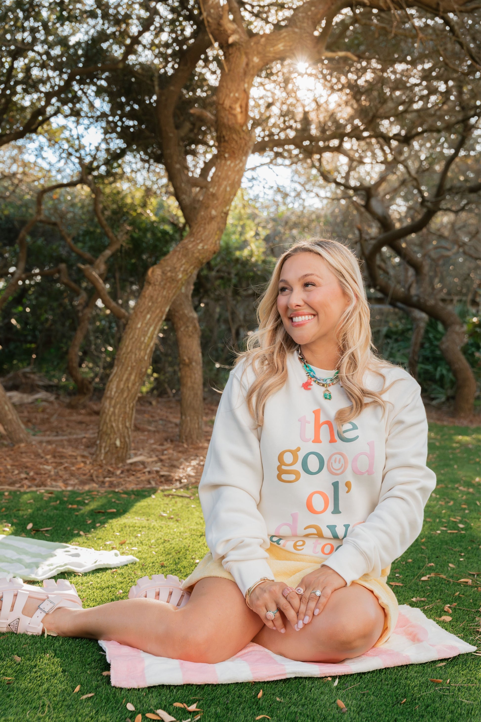 Woman sitting on a blanket outdoors wearing a Living Fully Co. sweatshirt with colorful text.