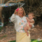 Woman holding a baby outdoors with trees and grass in the background.
