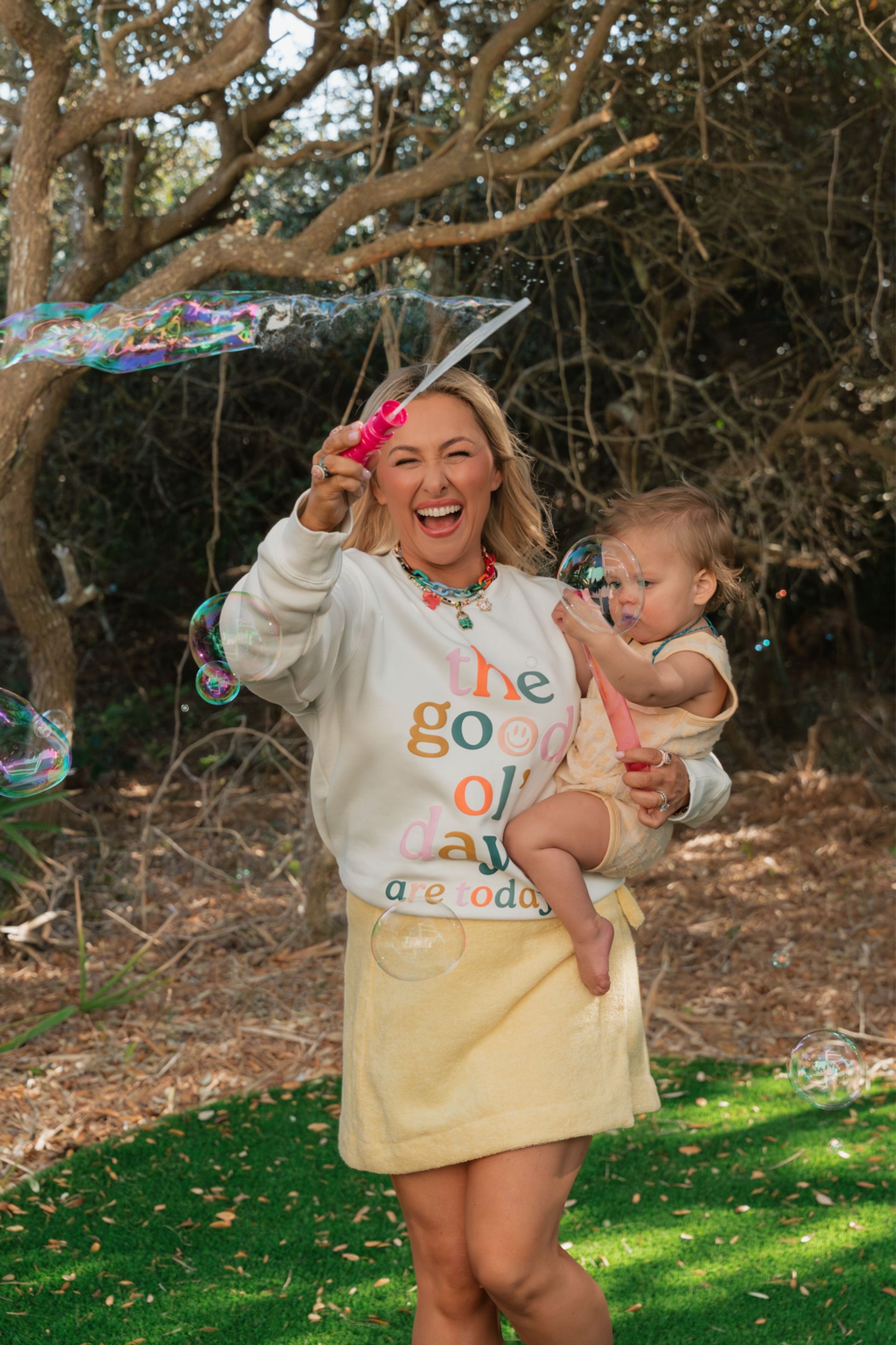 Woman holding a baby outdoors with trees and grass in the background.