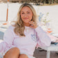 Woman in a pink sweatshirt with a blue oyster design sitting at a beachside table with flowers and a white umbrella.