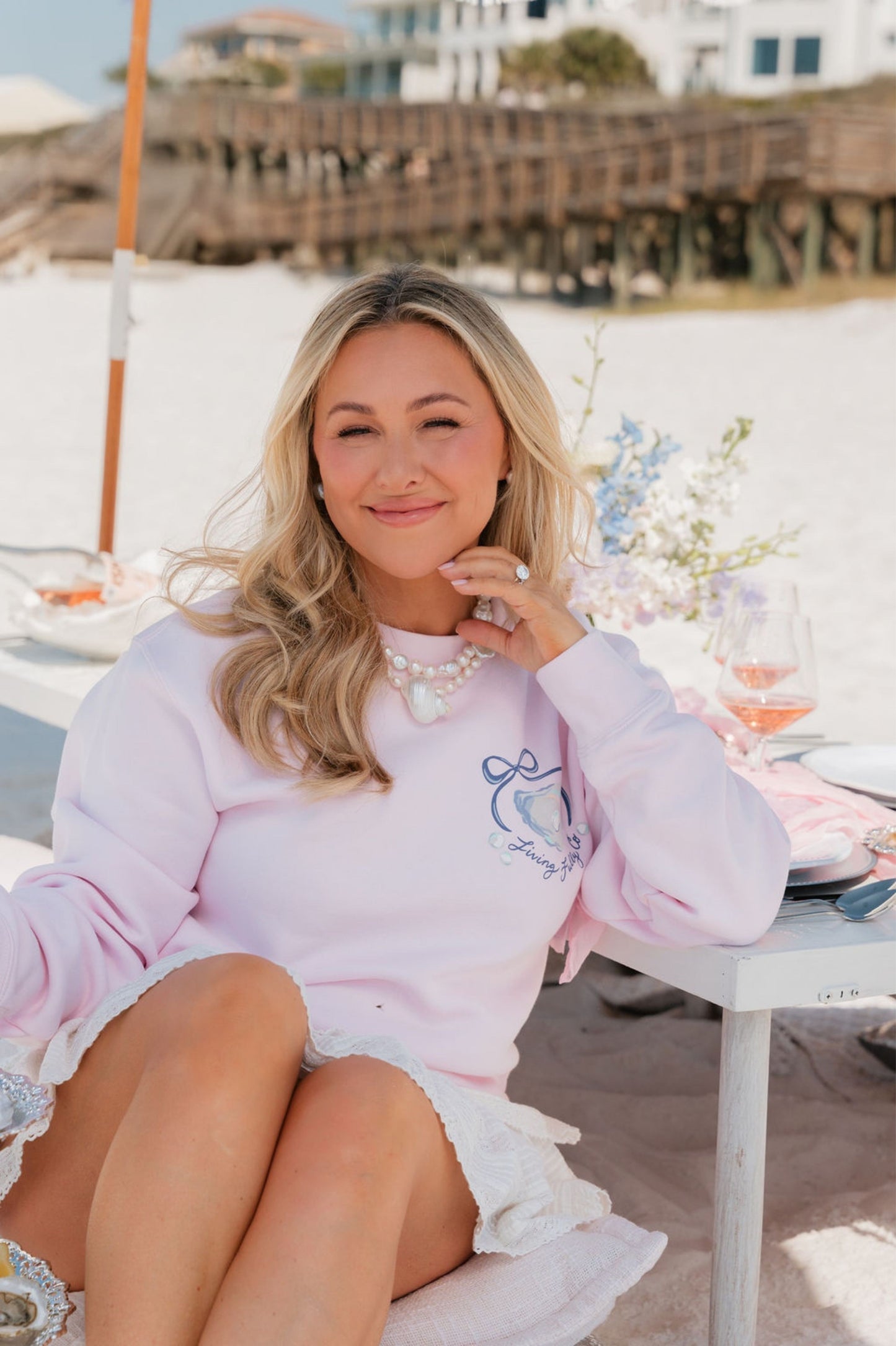 Woman in a pink sweatshirt with a blue oyster design sitting at a beachside table with flowers and a white umbrella.