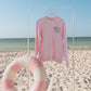 Pink long-sleeve sweatshirt on a hanger with a striped pink and white ring on a sandy beach.