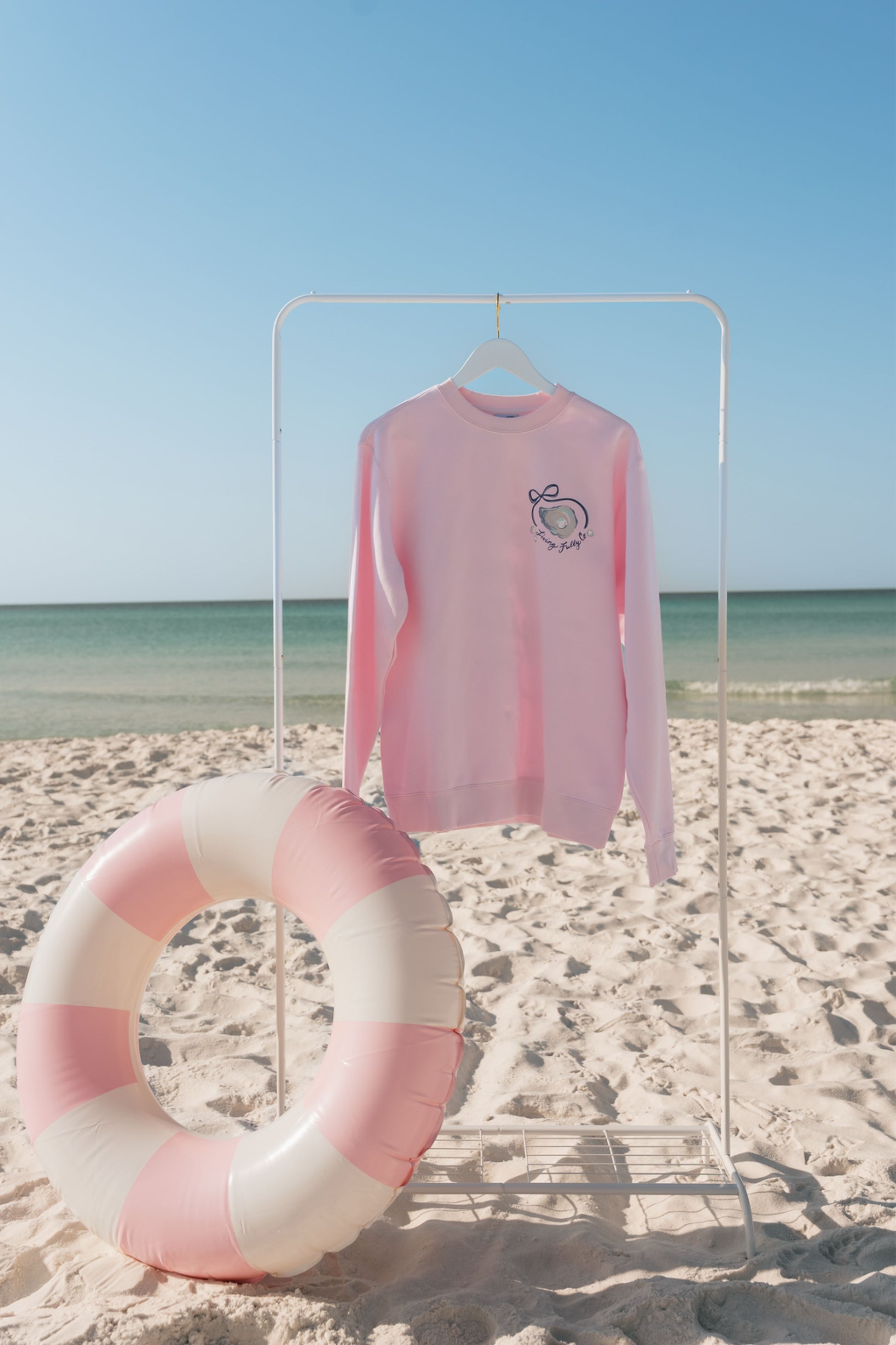 Pink long-sleeve sweatshirt on a hanger with a striped pink and white ring on a sandy beach.