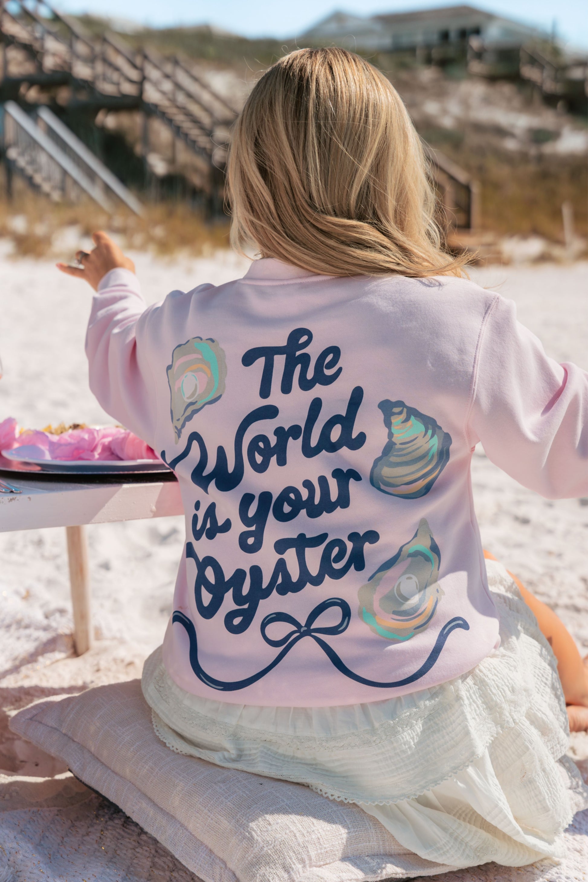 Person wearing a pink sweatshirt with 'The World is Your Oyster' text on a beach.