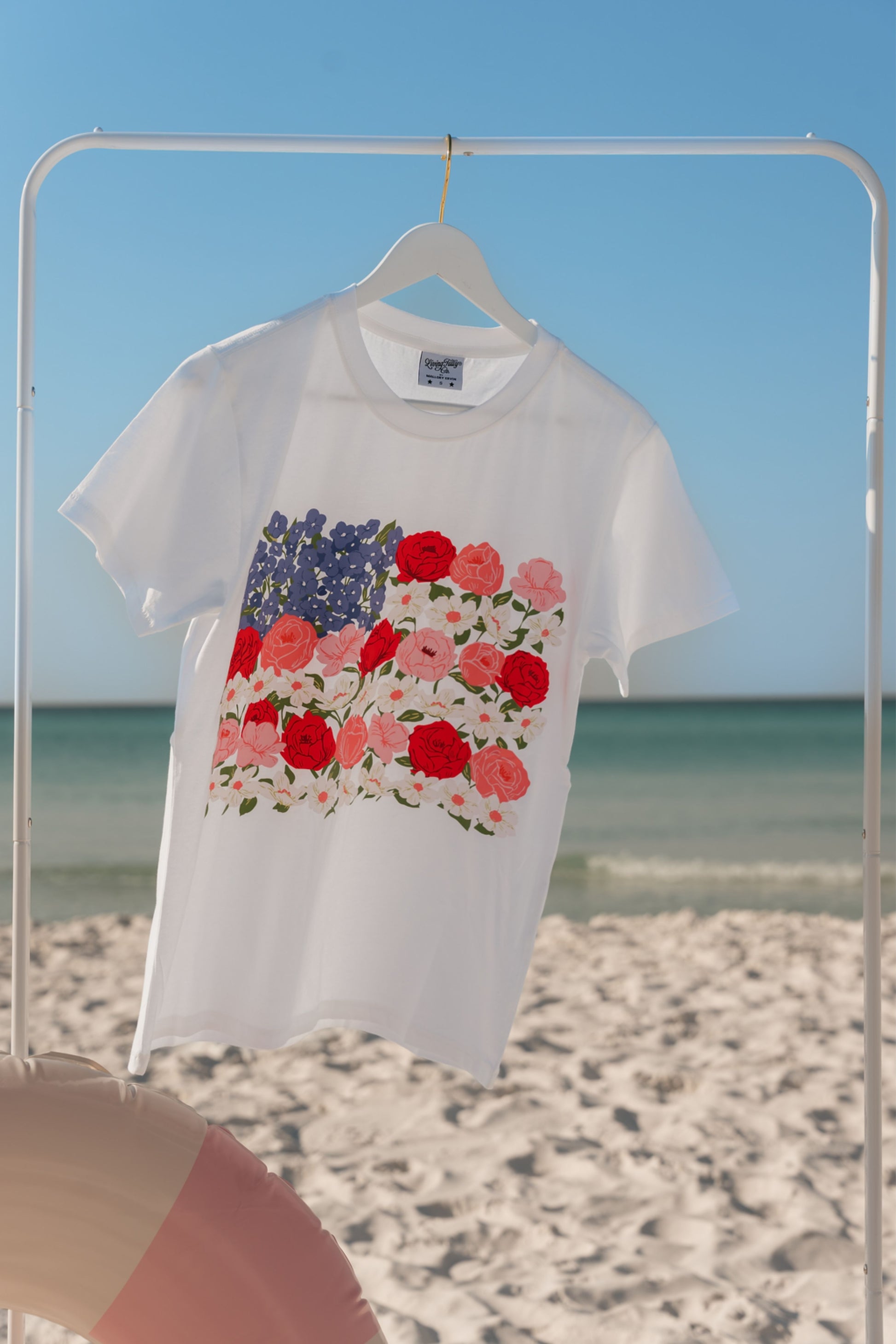 White t-shirt with floral design on a hanger against a beach background.