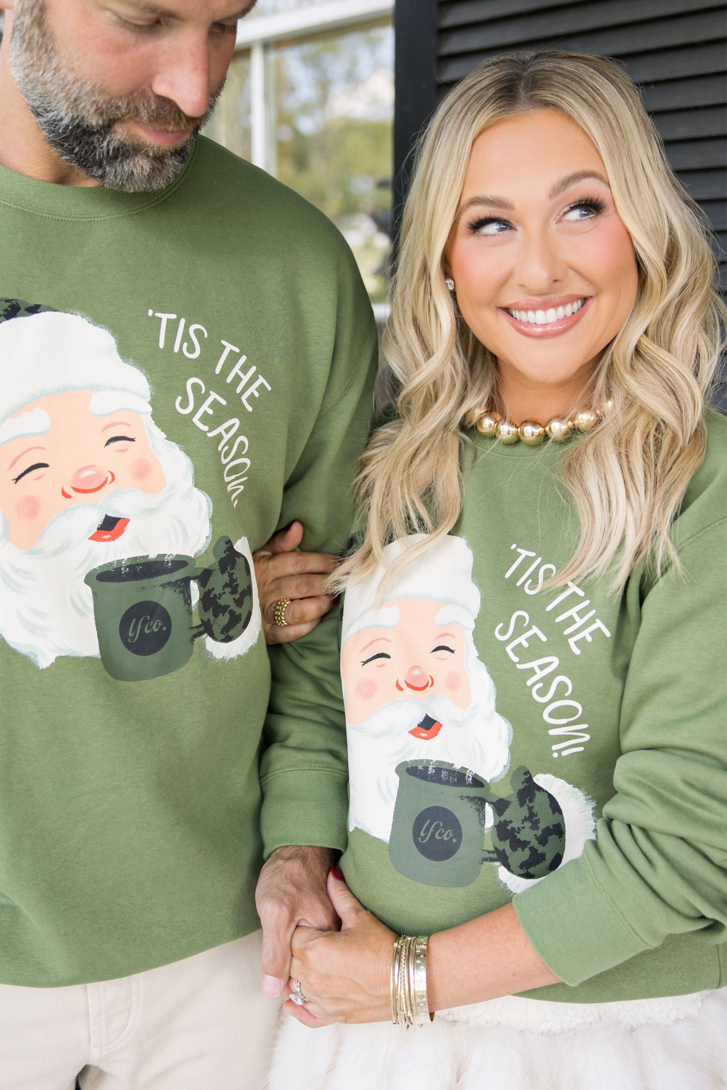 Two people wearing green sweaters with a Santa Claus graphic and 'Tis the Season' text.