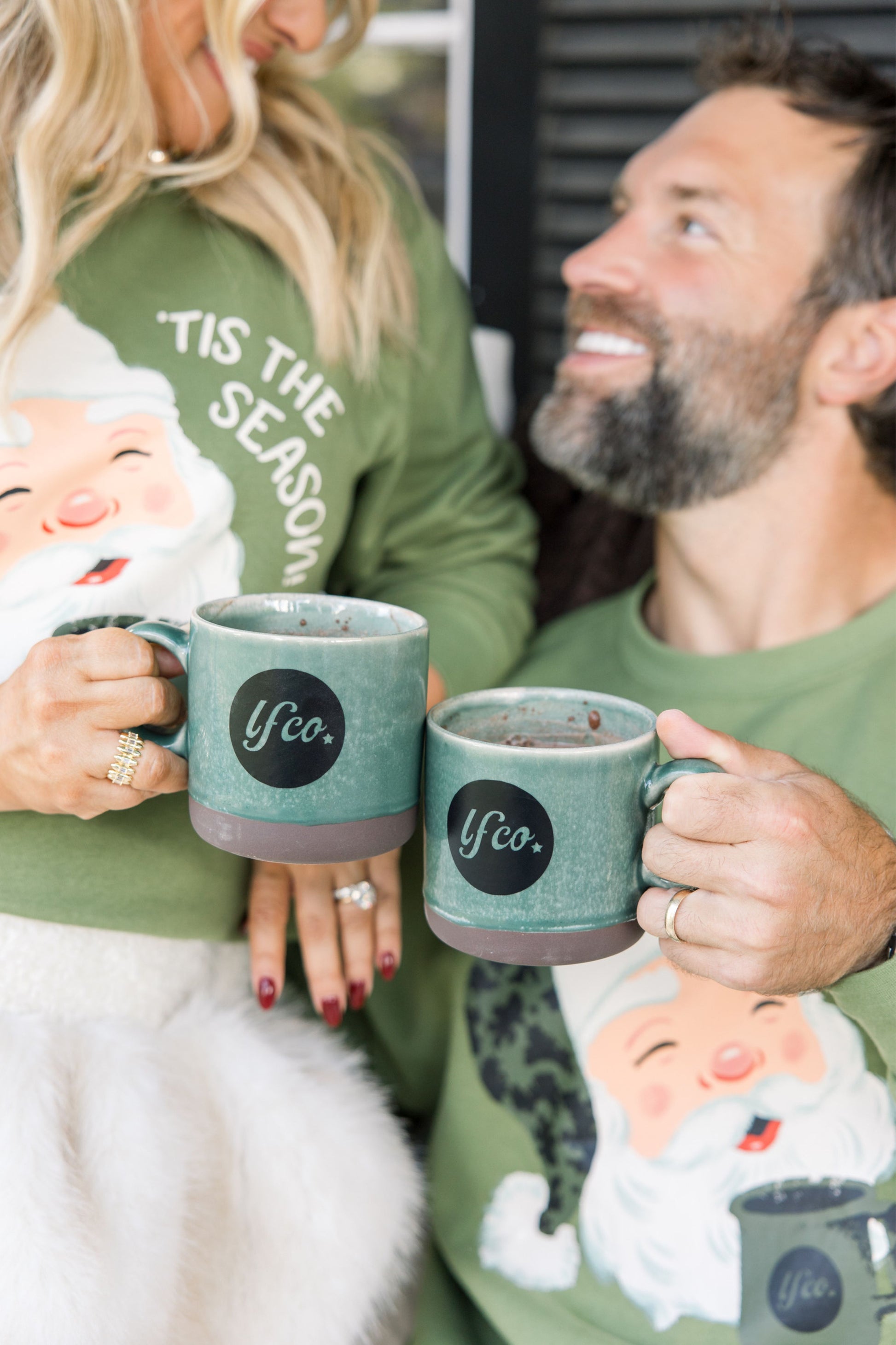 Two people holding mugs with a festive design, one person wearing a shirt with 'Tis the Season' text.