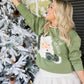 Woman in a green sweater with a Christmas design, standing next to a decorated tree.