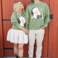 Two people wearing green sweaters with Santa Claus graphics in front of a red wooden door.