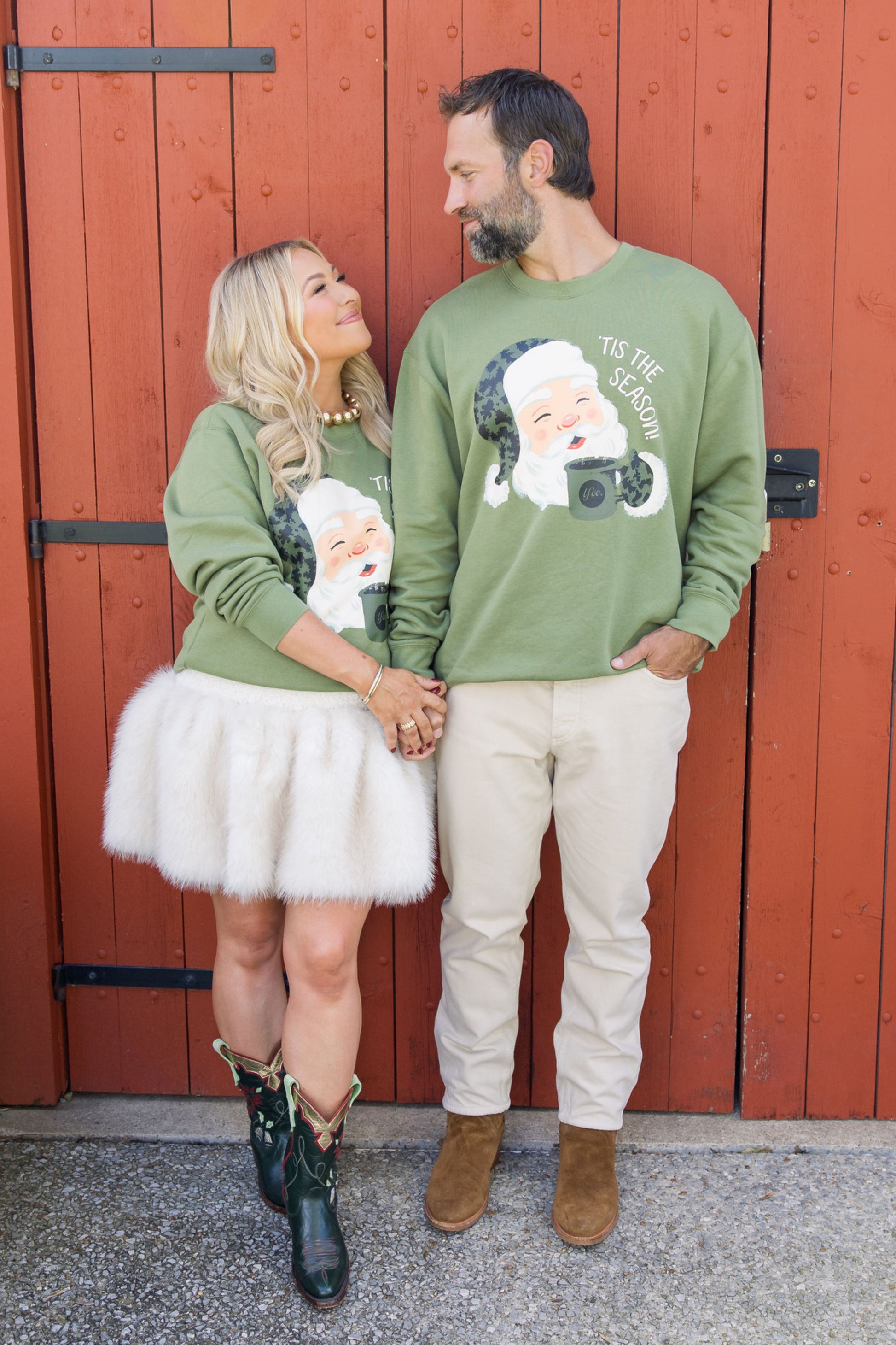 Two people wearing green sweaters with Santa Claus graphics in front of a red wooden door.