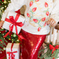 Person wearing a festive sweater with Christmas lights design and red sequin skirt, holding a gift in a decorated room.