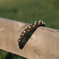 A dark brown fabric headband decorated with small white pearls, draped over a rustic wooden fence rail with a blurred green outdoor background.