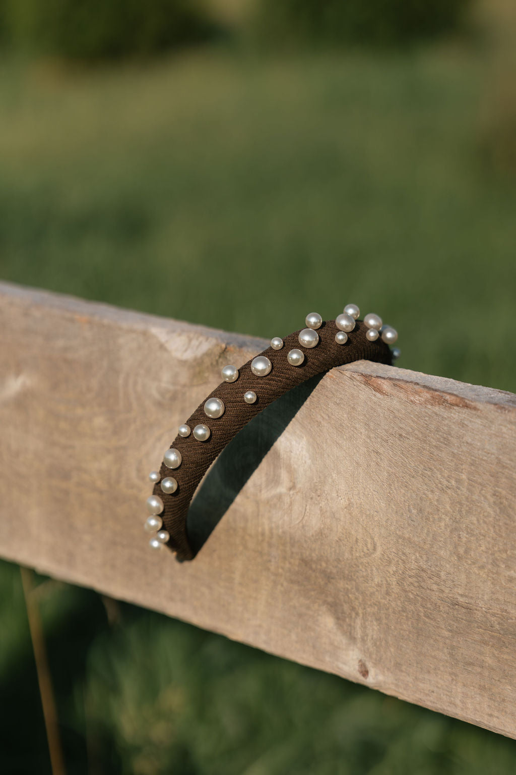 A dark brown fabric headband decorated with small white pearls, draped over a rustic wooden fence rail with a blurred green outdoor background.
