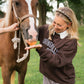 A woman in a brown sweatshirt and gingham shorts feeds a carrot to a brown horse outdoors on a grassy field. The woman wears a pearl headband, and the horse has a white blaze on its face.