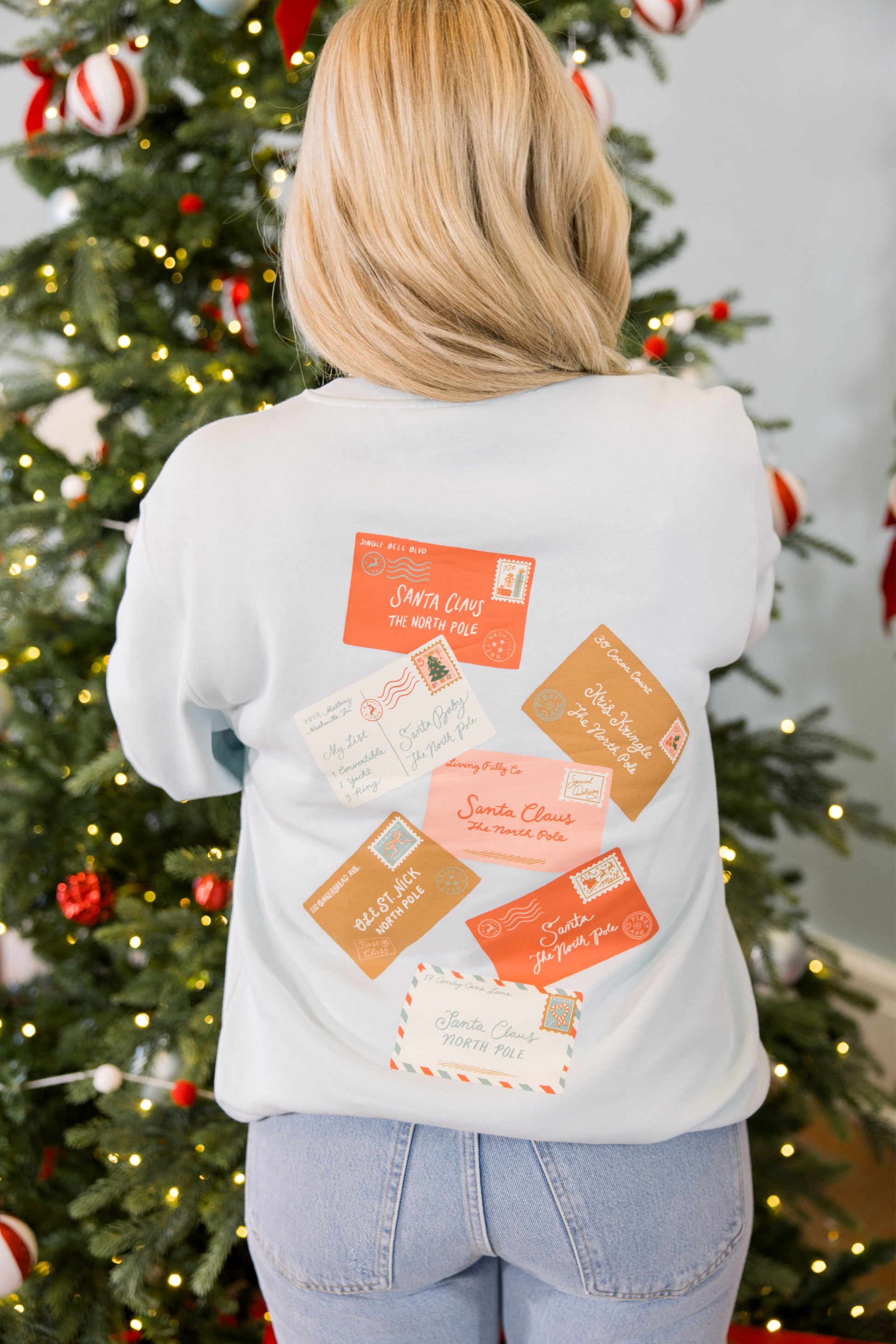 Person wearing a white sweatshirt with postal-themed designs in front of a decorated Christmas tree.