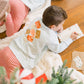 Child wearing a Christmas-themed sweatshirt with ornaments, sitting on the floor near a decorated Christmas tree.