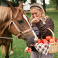 A woman in a brown sweatshirt and gingham shorts feeds an apple to a brown horse outdoors on a grassy field, holding a basket of red apples.