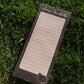 A rectangular notepad with lined writing space and a checklist on the left side, placed on green grass. The top reads 'Living Thankfully' and the bottom has a small decorative emblem.