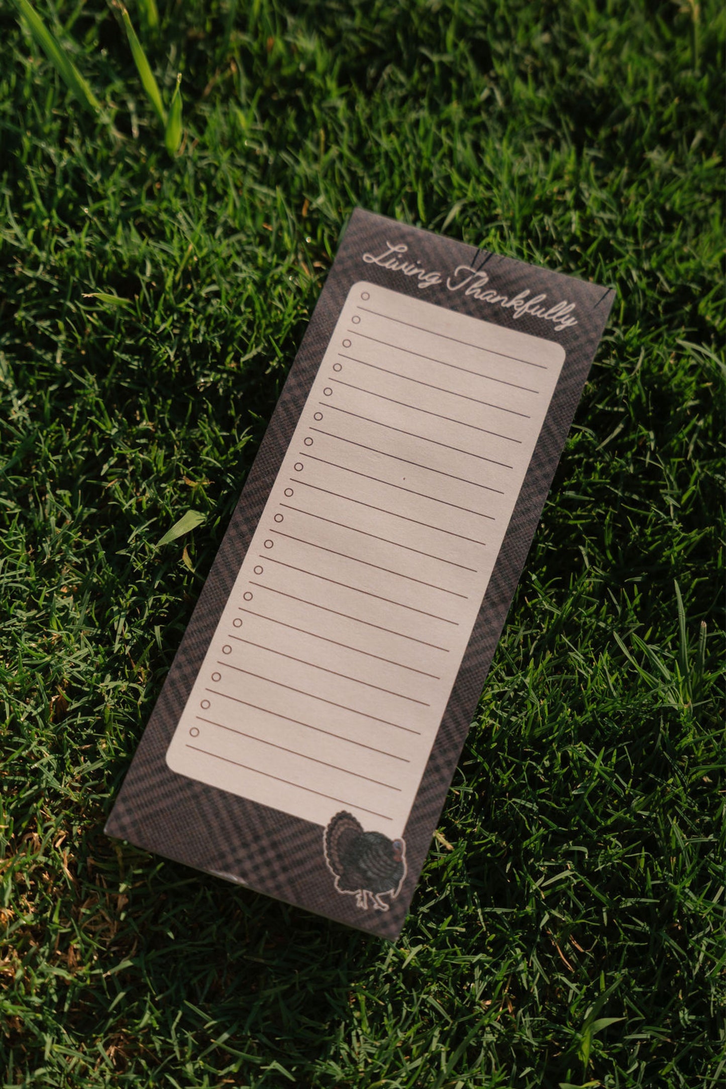 A rectangular notepad with lined writing space and a checklist on the left side, placed on green grass. The top reads 'Living Thankfully' and the bottom has a small decorative emblem.