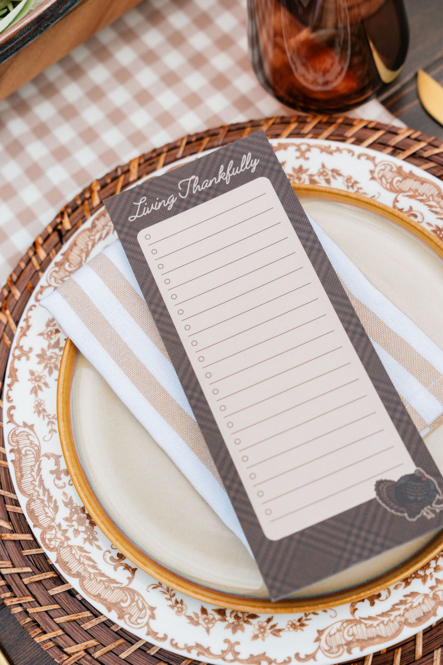 A rectangular 'Living Thankfully' gratitude card with lined writing space, styled on a beige and white striped napkin over a patterned ceramic plate and woven placemat.