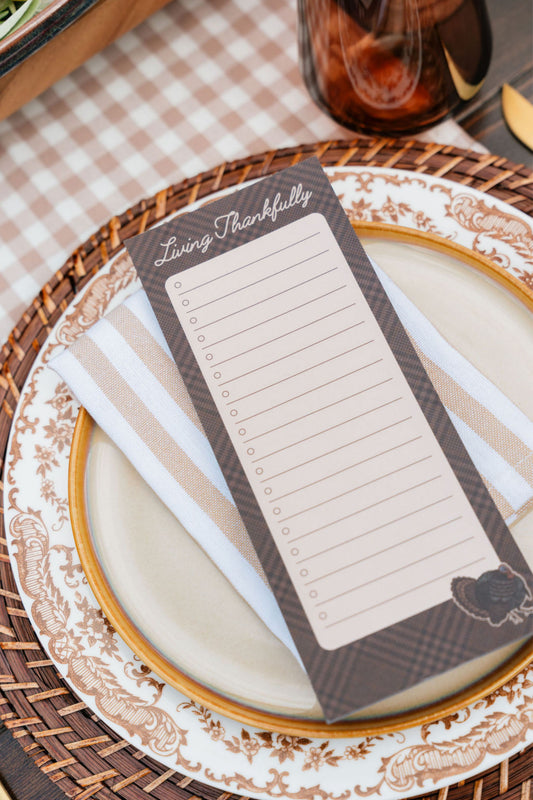 A rectangular 'Living Thankfully' gratitude card with lined writing space, styled on a beige and white striped napkin over a patterned ceramic plate and woven placemat.