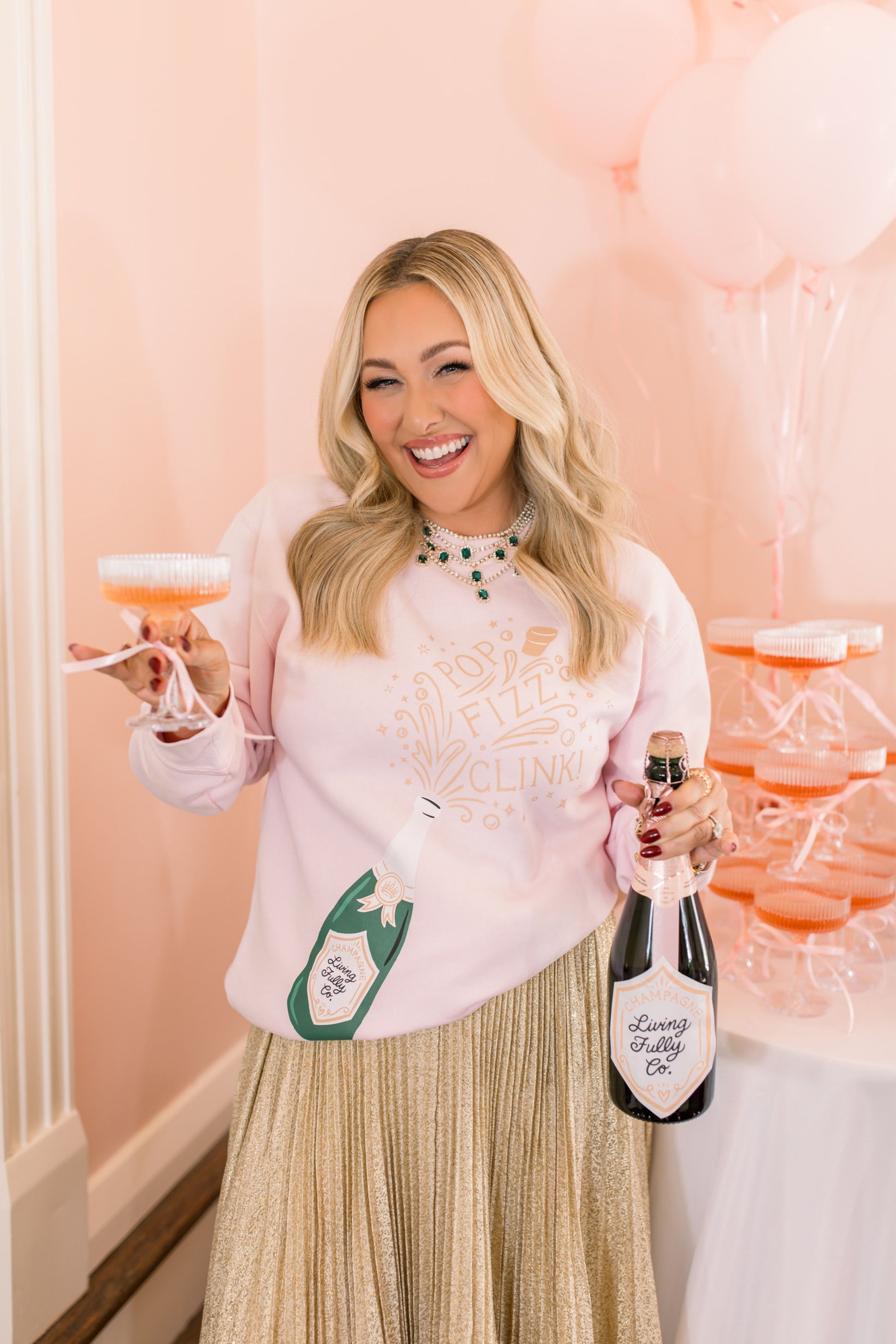 Woman holding a bottle of champagne and a cocktail in a celebratory setting with pink balloons.