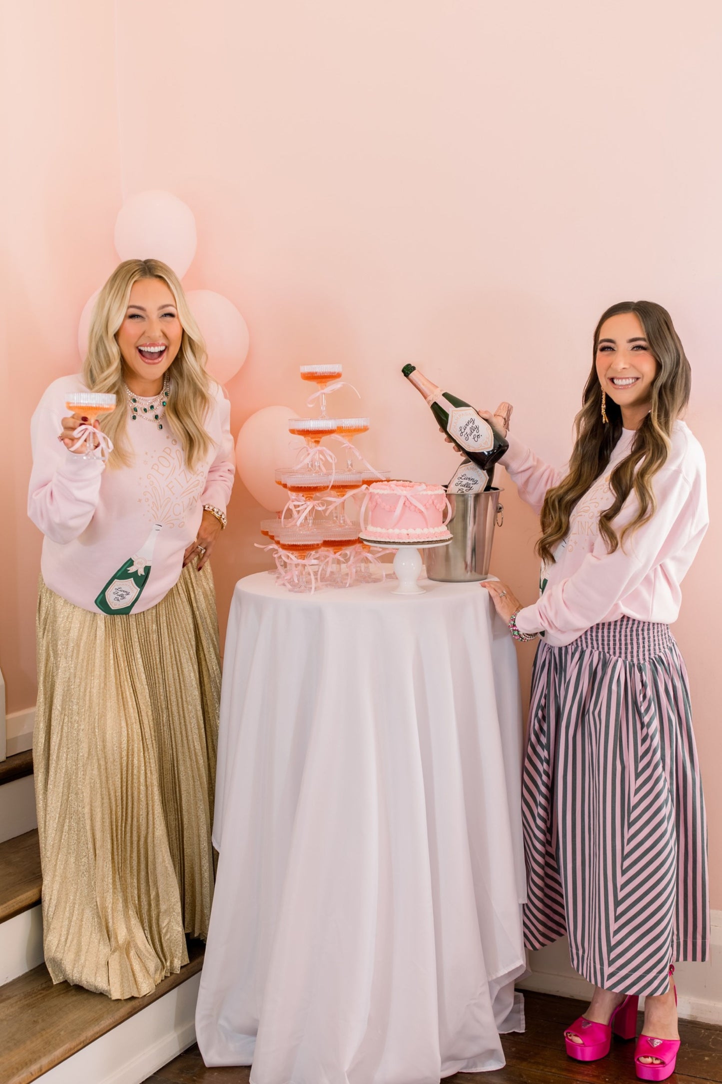 Two women celebrating with a cake and champagne bottle in a decorated room.