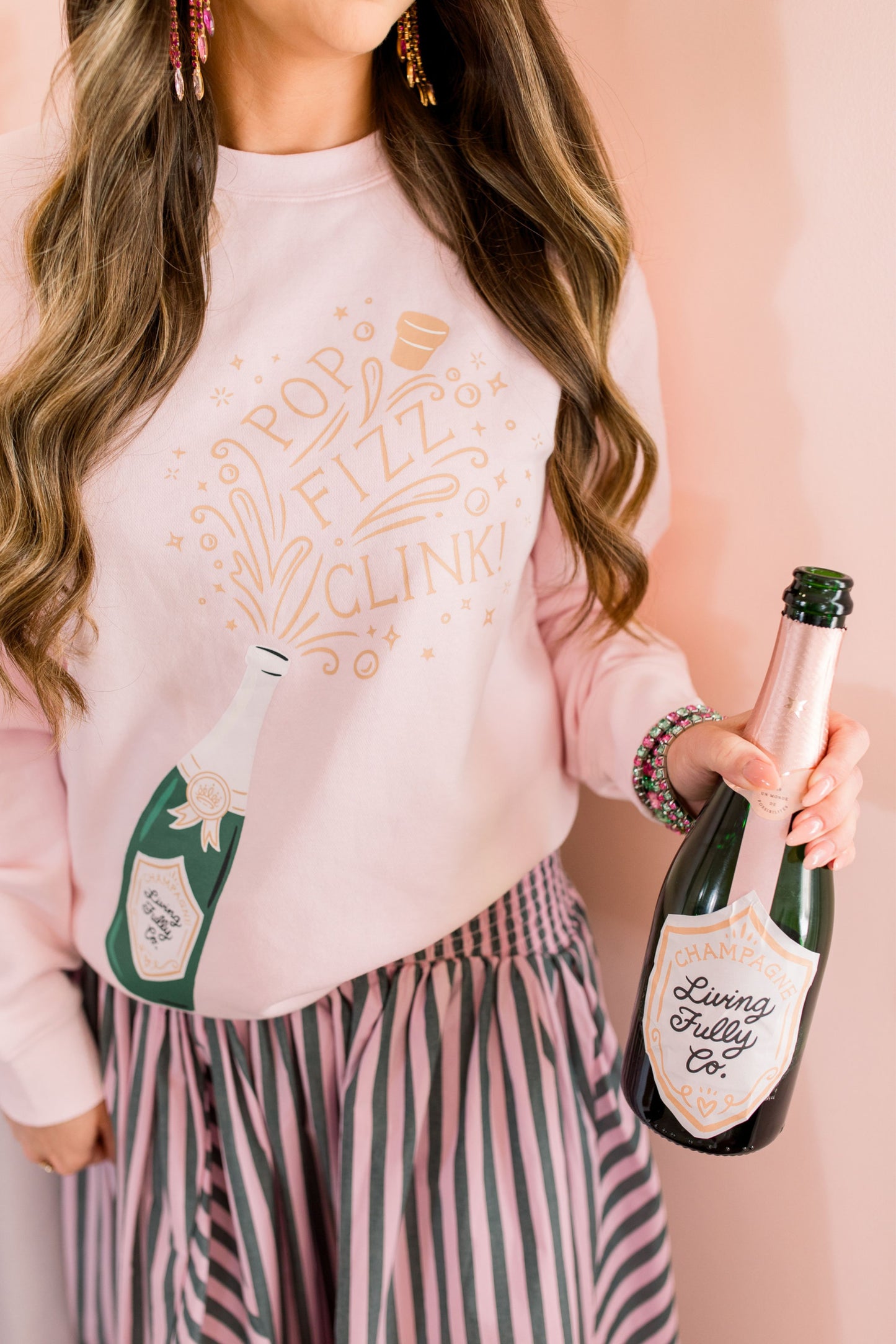Person wearing a pink sweatshirt with champagne bottle graphics and holding a real champagne bottle.